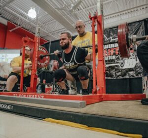 Personal trainer squatting with heavy weights at a competition, supported by spotters and surrounded by gym equipment.